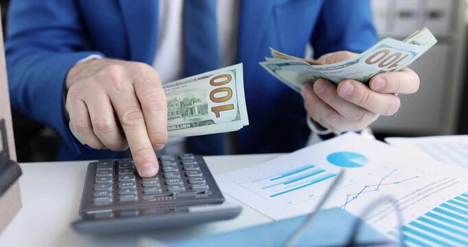 Businessman counts dollar bills over desk with calculator and info-charts. Man entrepreneur works with cash money conducting company audit in office
