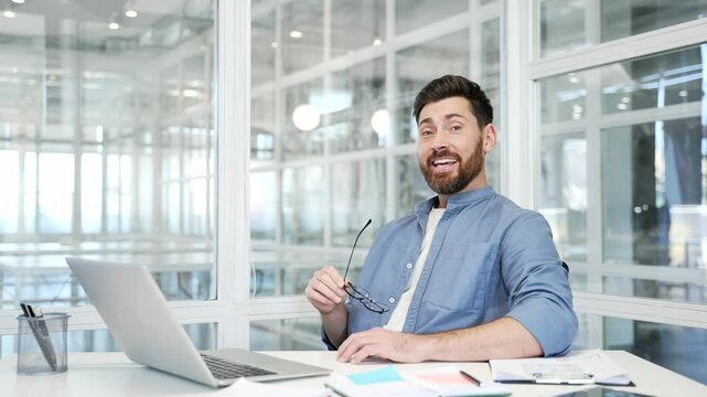 Happy businessman is satisfied with completed task on laptop while sitting at desk at workplace in a modern business office. Smiling worker successfully finished work on project and looking at camera
