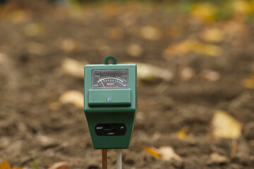 Close-up of a green soil moisture meter in a garden setting with blurred muddy background,