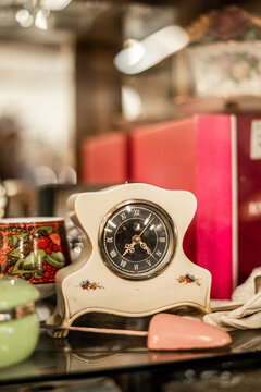 A vintage ceramic curved body mantel clock with a black clock face and Roman numerals, sitting on a display shelf surrounded by various antique items. 