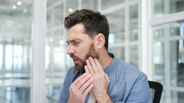 Upset businessman suffering from toothache while sitting at workplace at desk in business office. Male worker holds his hand on his cheek, massaging the sore spot. He needs to see a dentist. Close up