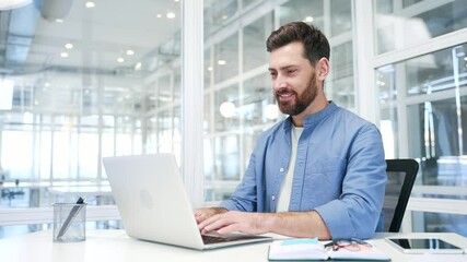 A smiling businessman working on a laptop sitting at a workplace in business office. Handsome bearded male worker typing on computer, banking, texting a client, chatting online or busy with a project - Powered by Adobe