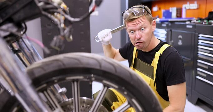 Funny dull mechanic scratches head back with wrench looking at motorcycle in car repair shop. Shocked worker doubted by luxury bike in car station