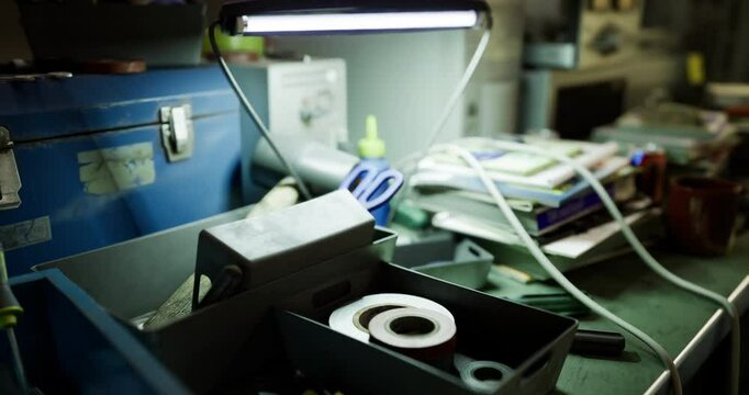 A workspace cluttered with tools, supplies, and a bright lamp. Scissors rest near containers. The setting appears busy, ideal for creative projects or repairs.