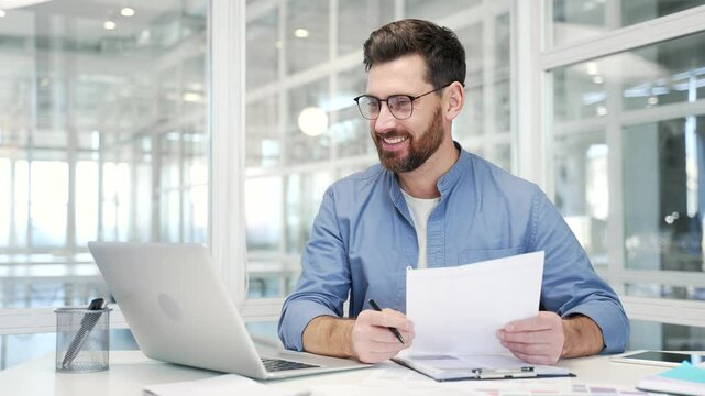 Joyful businessman is satisfied with the results of a financial report holding documents in his hands sitting at workplace in a business office. Entrepreneur or owner is happy with positive indicators
