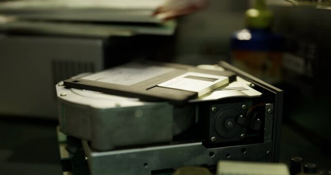 A floppy disk rests on a classic hard drive in a cluttered tech workspace. Surrounding tools and machinery hint at an ongoing electronics project. The setting indicates a vintage technology focus.