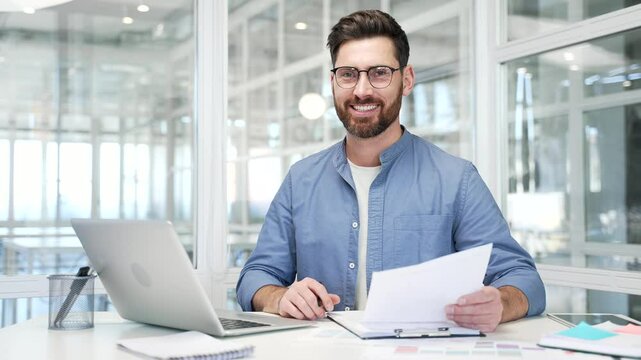 Portrait of confident businessman sitting at desk at workplace in modern business office. Successful handsome smiling entrepreneur financier busy with paperwork working on laptop and looking at camera
