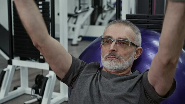 Caucasian bearded mature man with glasses wearing grey t-shirt performing front raises on cable machine leaning on fitness ball with his back during workout in rehabilitation centre
