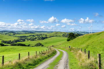 scenic rural landscape featuring winding dirt road, lush green hills, and wind turbines under bright blue sky