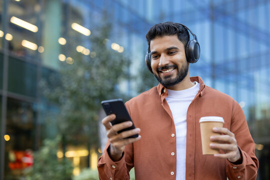 An attractive man listens to music while scrolling on his phone and holding a coffee cup. The background shows a modern building.