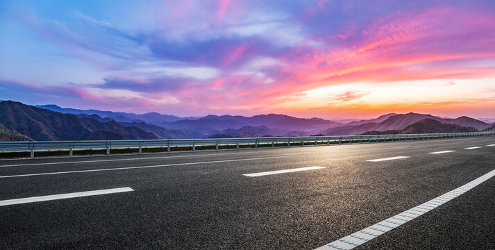 Empty asphalt highway leads towards stunning mountain range at colorful sunset