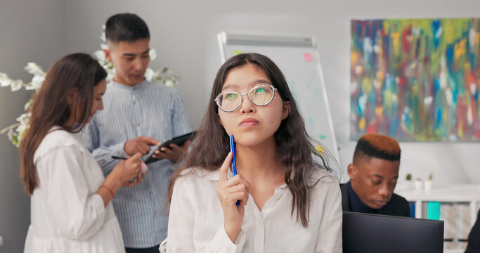 In the company office, a brunette with glasses sits at her desk in a plaid skirt and white shirt. She holds a notebook in one hand, with a pen resting on her cheek, lost in deep thought, searching for