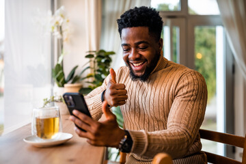 A young black man sits at the bar, casually dressed. He is using smartphone and having video call.