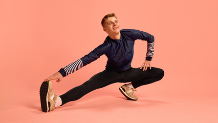 Sporty man smiling while stretching legs sideways, reaching for foot on pink background, wearing...