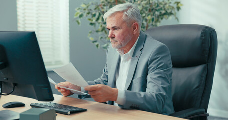 Mature, gray-haired, elegant businessman sits in office at the computer, completes company...