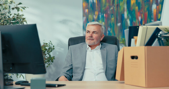 An elderly man with gray hair sits on a chair at an office desk, next to him a box of packed things, a guy in a suit looks to the side as he recalls years of working for the company he is retiring