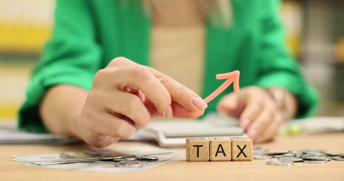 Woman in jacket placing small arrow on top of wooden blocks with word Tax. Coins scattered around table in office suggesting financial matters