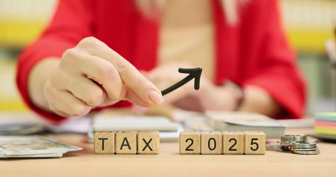 Female worker placing dark arrow pointing upwards on top of wooden blocks with phrase Tax 2025. Stationery scattered around table in office room