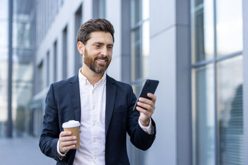 Successful businessman walking through the city with phone in hand, smiling happily. Man in business suit browsing social media and writing text message, with cup of hot drink in hand.
