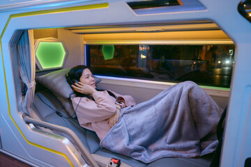 Asian woman listening to music on a night sleeping bus while relaxing with blanket.