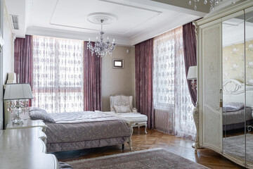 design of bedroom in retro style. interior with retro furniture. bedroom is decorated in beige and purple shades. double bed, wardrobe, chest of drawers, chair, mirror, windows with purple curtains.