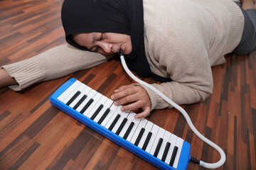 Asian muslim woman wearing hijab playing pianica while lying on floor, melodica blow organ