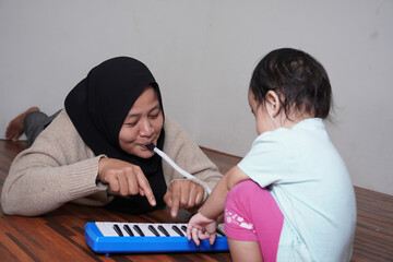 Asian muslim mother wearing hijab playing pianica with her baby daughter, melodica blow organ