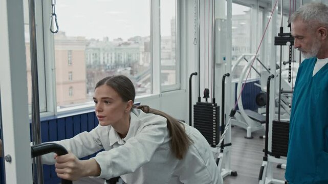 Young woman doing cable kickbacks when mature male physiotherapist helping and controlling her in space equipped with machines for rehabilitation exercises