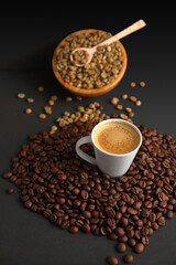 Cup of espresso coffee surrounded by toasted coffee beans and a wooden bowl in the background with green coffee beans