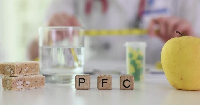 Person holds measuring tape at desk with wooden blocks showing PFC assessing dietary balance. Protein bars yellow apple and glass of water on table