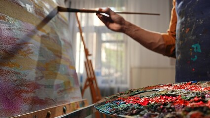 Obraz premium Person in apron stands at easel and drawing on canvas with paintbrush and palette during art class in studio, close up shot.