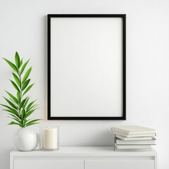 White minimalist interior scene featuring a blank black frame, a potted plant, a candle, and a stack of books on a white dresser against a textured white wall. 