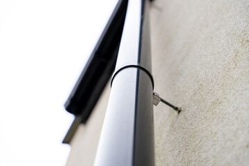 Close-up view of black downspout pipe attached to a house wall with white facade and mounting...