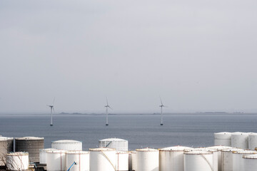 Offshore wind turbines aligned with coastal oil tanks, visualizing contrast between fossil energy and renewable development in global energy economy.