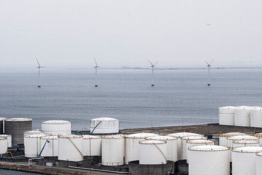 Industrial storage tanks aligned by the coast with offshore wind turbines in the background, combining energy production and maritime infrastructure.