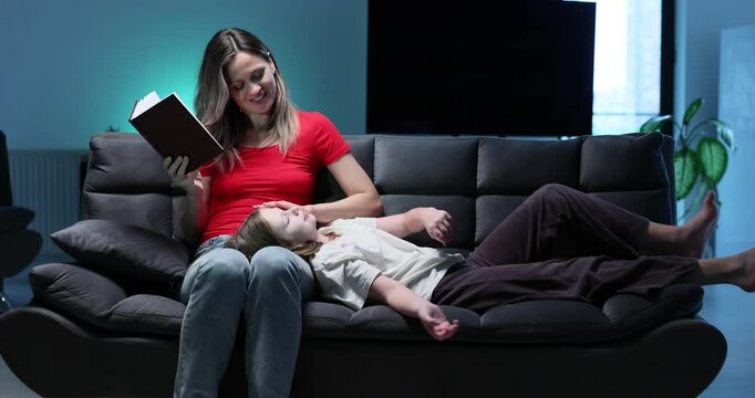 Loving mother holds open book while daughter lies cozy on laps in living room. Woman reads bedtime story to cute girl sitting on sofa in evening