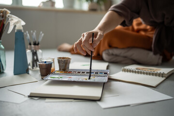 Unrecognizable woman artist hand mixing watercolors for sketch in notebook on floor in studio....