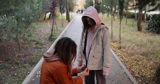 Woman helps young girl with jacket in cold weather in autumn. Moment of assistance and warmth between mother and daughter near trees with pathway