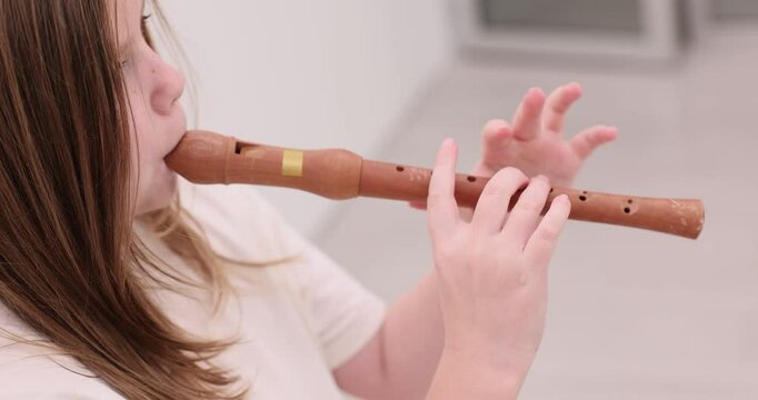Focused schoolgirl with long hair plays wooden recorder in light studio room. Active preparation rehearsal of child before music performance on flute