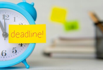 A light blue alarm clock and a sticky note with blurred stacks of books in the background. Deadline, reminder, urgent concept.