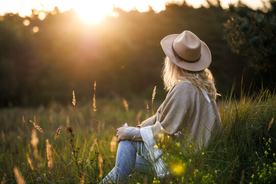 Woman with poncho and hat spending time in nature for mental wellbeing and improve mindfulness. Relaxation on meadow during sunset