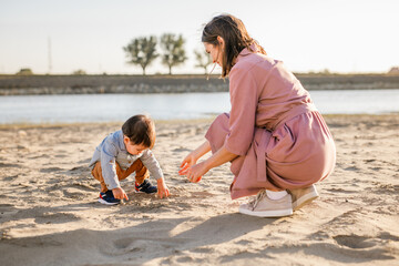 Young woman in a pink dress playing in the sand with her one-year-old son