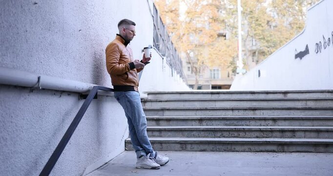 Man with smartphone and coffee leans on railing standing on street underpass staircase. Worried guy looks back to check clothes cleanness outdoors