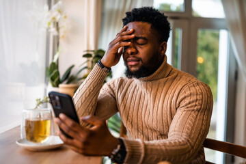 A young black man sits at the bar, using smartphone, messaging online. He is anxious and preoccupied.