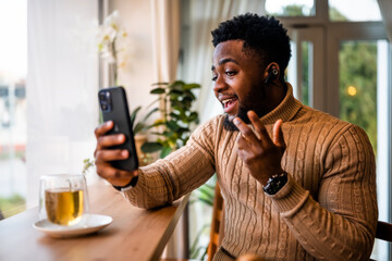 A young black man sits at the bar, casually dressed. He is using smartphone and having video call.