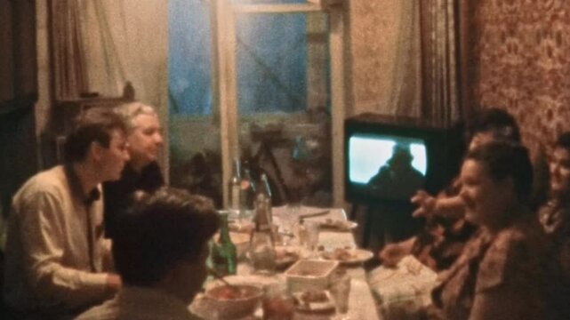 Large family having dinner in TV front at home indoor. Many people, smiling friends chatting, eating at dinner table together. Happy generation meeting. Vintage film. Retro archive. 1980s Ukraine 