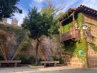 A peaceful courtyard featuring a rustic ivy-covered house, stone benches, lush trees, colorful yarn art installation, and playful recycling bins.