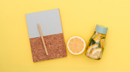 Eco-material notebook with a zesty lemonade bottle and juicy orange set against a cheerful yellow backdrop, capturing the essence of eco-conscious living and summer freshness in a top view flat lay