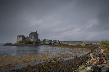 Eilean Donan Castle stands majestically amid autumn hues, its reflection shimmering in calm waters. Misty Highlands and golden trees create a scene of timeless beauty.