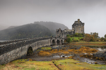 Eilean Donan Castle stands majestically amid autumn hues, its reflection shimmering in calm waters. Misty Highlands and golden trees create a scene of timeless beauty.